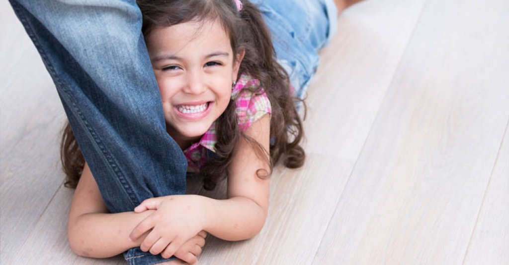 A girl hugging her parent's leg on a wood floor.
